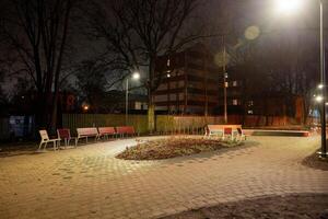 A small urban park at night with a curved pathway, red and white benches, sparse vegetation, and multi story buildings in the background. photo