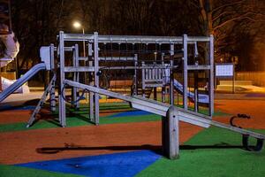 Nighttime view of a playground featuring a wooden play structure, slides, climbing nets, and seesaw. Colorful flooring and bare trees are visible. photo