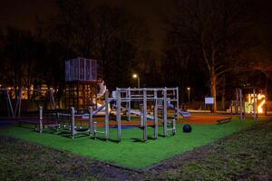 A well lit playground at night featuring a cube like climbing structure, slides, and colorful rubberized flooring, surrounded by bare trees and streetlights. photo