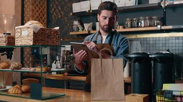Smiling Caucasian guy delivery barista man calculate order price bill on tablet tech small business in cafeteria store catering restaurant waiter worker male give package food for client cafe service video