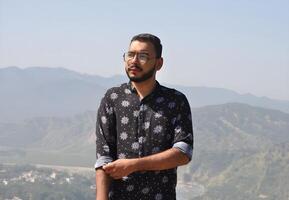Handsome bearded Indian man wearing a blue shirt and glasses, standing triumphantly on top of a mountain with a breathtaking view photo
