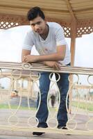 Young Asian man leaning on the railing of a park bench, gazing directly at the camera with a friendly expression photo