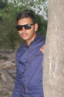 Young man wearing sunglasses, leaning casually against a tree and looking directly at the camera with a relaxed expression photo