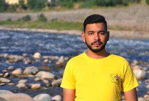 young man with a stylish beard in a yellow t-shirt standing against a blurred mountain river background photo
