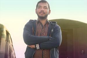Portrait of a young Asian man standing with arms crossed confidently, set against the backdrop of a train photo