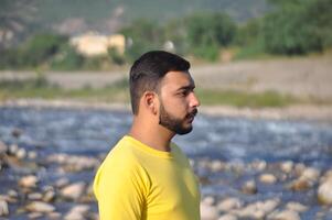 Handsome man with a beard in a yellow t-shirt standing against a blurred mountain river background photo