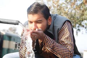 Young man drinking water from a public fountain in a park, with a street and greenery in the background photo