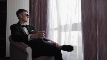 Groom in tuxedo relaxing by a window with a drink, groom sitting in a chair by a window, holding a drink, in formal attire. video