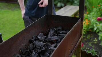Experienced cook preparing charcoal grill in sunny garden, rusty barbecue surrounded by wooden bench and blooming flowers, capturing summer leisure moment video