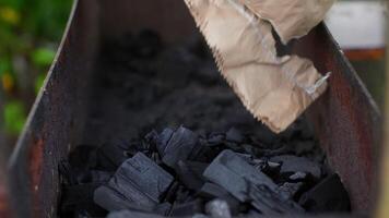 Black charcoal pouring from crumpled brown paper bag into rusty metal grill, creating texture and preparing outdoor cooking surface for upcoming barbecue gathering video