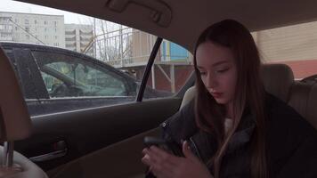 Young woman passenger using her smartphone while sitting in the backseat of a car on a winter day, with snowy buildings visible through the window video