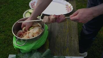 Cook stringing pieces of raw pork meat onto metal skewers outdoors on a wooden table, with a pot of marinated meat and sliced zucchini nearby video
