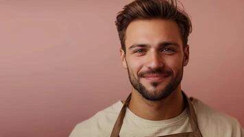 Smiling young man with a beard and wavy brown hair, wearing a beige apron, looking friendly and confident against a soft pink background video