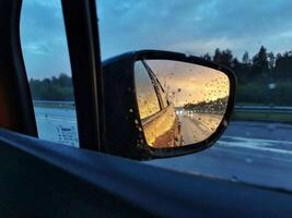 Raindrops on the car side mirror reflect a tranquil sunset on a forest road during evening travel photo