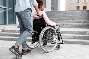 Young black guy pushing wheelchair with handicapped woman, having problem entering building, standing in front of stairs without ramp. Problems with accessible environment for disabled people photo