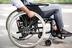 Unrecognizable young black man in wheelchair going for walk in city park, closeup of legs. Cropped view of millennial African American guy with physical disability spending time outdoors photo