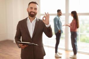 Successful Deal, House Ownership Concept. Portrait Of Smiling Professional Real Estate Agent In Suit Showing Okay Sign Gesture, Selective Focus. Buyers Talking In Blurred Background photo