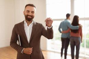 Portrait Of Cheerful Professional Real Estate Agent Holding Clipboard And Showing Keys In Hand, Selective Focus. Young Newlyweds Choosing New Home Looking Out Window Hugging In Blurred Background photo