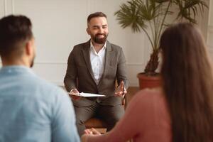 Smiling therapist talking with young millennial patients, back view of happy married couple spouses sitting on sofa holding hands visiting professional family counselor, resolving issues photo