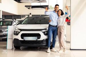 Joyful middle-eastern couple standing next to luxury white car and hugging at dealership automobile salon, rasising hands up, showing key from their new auto, cheerfully smiling at camera, full length photo