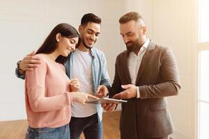 Portrait Of Smiling House Consultant In Suit Showing Modern Apartment To Young Arab Clients, Pointing At Digital Tablet With Project, Discussing House Plan, Selling Flat To Happy Middle Eastern Family photo