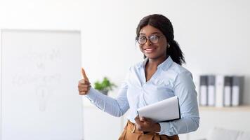 Psychology teacher or business coach giving seminar or presentation, showing thumb up gesture at office. Black female lecturer recommending training course on personal growth and motivation photo