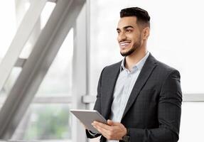 Portrait Of Handsome Middle Eastern Businessman Using Digital Tablet While Waiting For Flight At Airport Terminal, Smiling Arab Man In Suit Holding Tab Computer And Looking Away, Copy Space photo