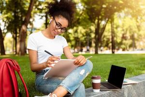 Black Student Girl Sketching Or Taking Notes Learning Online Sitting With Laptop Computer In University Campus Park Outside, Preparing Lesson. Education And E-Learning photo