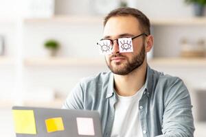 Tired millennial guy having stickers with drawings of open eyes on his glasses, sleeping in front of laptop at home office. Exhausted young man overworking, falling asleep a workplace photo