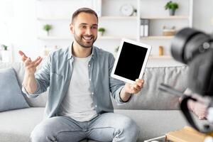 Happy male influencer filming review of modern digital tablet on dslr camera at home, mockup on screen. Cheerful Caucasian blogger advertising modern tech product, streaming live on web photo