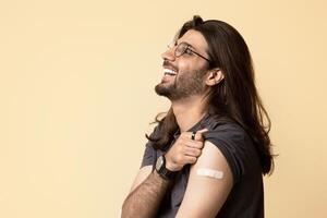 Positive middle-eastern long-haired young man in casual sitting on chair over studio background, got vaccinated against coronavirus, showing plaster, looking at copy space and smiling, panorama photo
