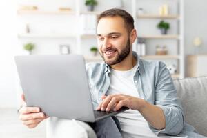 Cheerful millennial man sitting on couch with laptop computer, working or studying online from home office. Joyful Caucasian guy participating in web lecture, taking part in conference photo