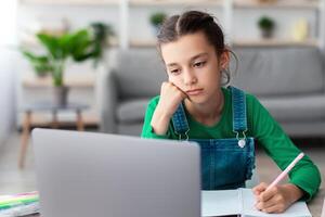 Too Tired For School. Portrait of bored upset student girl sitting at desk, doing homework, writing in her notebook and looking at laptop screen, studying online, selective focus. Home Based Learning photo