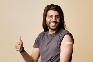 Cheerful long-haired indian guy showing thumb up, smiling at camera, got vaccinated against COVID-19, having adhesive medical band on shoulder, recommending immunization, studio background, copy space photo