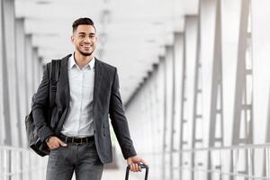 Portrait of young middle eastern man walking in airport terminal with luggage, smiling arab guy carrying suitcase while going to boarding gate, enjoying air travels and business trips, copy space photo