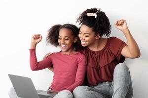 Emotional african american mom and kid gaming online together, using laptop, sitting on floor by white empty wall, raising hands up and smiling, playing games, panorama with copy space photo