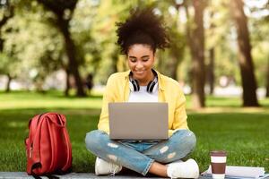 E-Learning. African American College Girl Using Laptop Having Distant Online Lecture Sitting On Grass In Park Outdoors. Female Student Browsing Internet Via Computer Learning Remotely photo