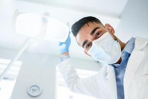 Low Angle View Of Arab Dentist Doctor Turning On Lamp Before Check Up With Patient, Professional Middle Eastern Man Stomatologist Wearing Medical Face Mask Looking At Camera, Closeup Shot photo