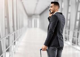 After Arrival. Smiling Arab Businessman Talking On Cellphone While Passing Airport Gate, Cheerful Middle Eastern Man Walking With Suitcase In Terminal And Enjoying Mobile Call, Rear View photo