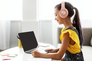 Side view profile portrait of girl in pink headset doing homework, sitting on couch at table using laptop with empty screen, writing essay or making test, taking notes in textbook, free copy space photo