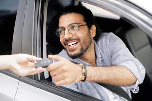 Handsome middle-eastern young man sitting inside nice automobile and taking car key from leasing or renting service person. Cheerful arab guy doing test-drive before buying new automobile photo
