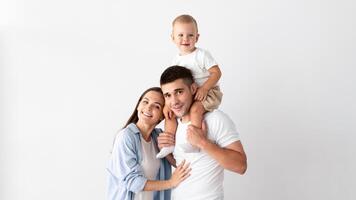 Portrait Of Happy Young Mother, Father And Their Cute Toddler Son Posing Together Over White Wall At Home, Cheerful Parents And Adorable Infant Child Embracing And Smiling At Camera, Copy Space photo