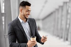 Young Arab Man Using Mobile Phone And Drinking Coffee While Waiting For Flight In Airport, Smiling Middle Eastern Guy Standing In Terminal, Messaging On Smartphone And Enjoying Takeaway Drink photo