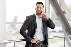 Handsome Middle Eastern Businessman Talking On Cellphone While Waiting At Airport, Smiling Arab Man Standing Near Window At Terminal And Enjoying Pleasant Mobile Call Conversation, Free Space photo