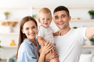 Happy Young Family With Little Infant Son Taking Selfie Together At Home, Cheerful Parents And Their Cute Toddler Child Posing For Photo In Domestic Interior, Looking And Smiling At Camera, Closeup