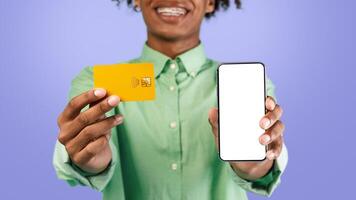 Cool mobile shopping app. Excited young black guy holding credit card and showing cellphone with blank screen to camera. Gadget with copy space for mockup, digital pay photo