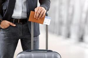 Ready For Trip. Unrecognizable Man Standing With Suitcase, Passport And Tickets At Airport Terminal, Male Traveller Waiting For Air Flight Near Boarding Gate, Cropped Image, Closeup Shot photo
