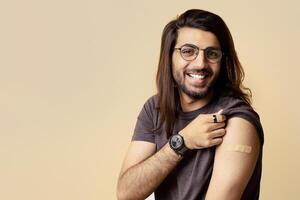 Closeup portrait of cheerful indian guy showing medical band on his shoulder after vaccination and smiling at camera, beige studio background, copy space. Immunization during coronavirus pandemic photo
