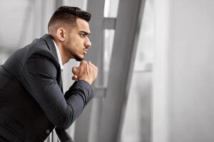 Portrait Of Thoughtful Arab Businessman Looking From Window While Waiting At Airport, Pensive Middle Eastern Man Leaning On Handrail And Touching Chin, Thinking About Something, Side View photo
