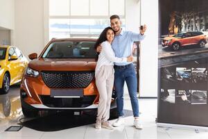 Happy middle eastern couple young man and woman standing by nice brand new car, holding key, embracing and smiling, buying new auto together at luxury showroom, panorama with copy space photo